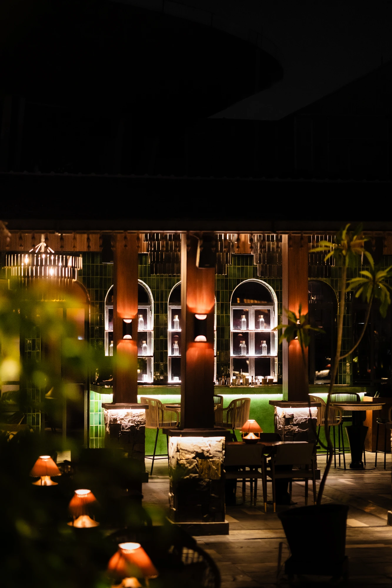 A view through greenery and timber pillars to the glowing green bar and lamp-lit seating at Upstairs.