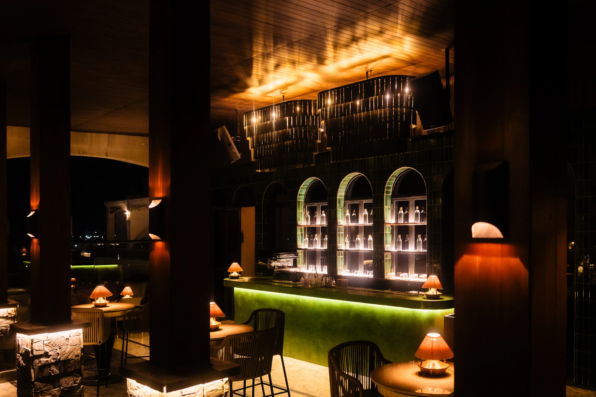 The Upstairs bar with green arched shelving, amber table lamps and stone-clad timber columns.